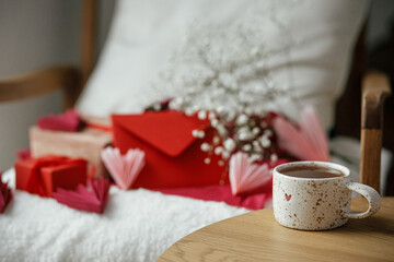 Cute stylish cup of tea with heart on wooden table against modern armchair with gifts, red envelope and white flowers. Valentine morning surprise for beloved. Happy valentines day!