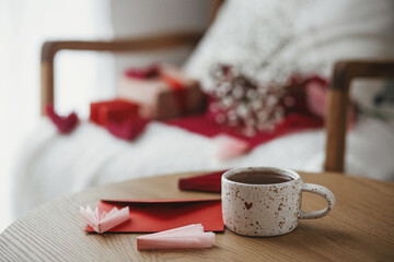 Cute stylish cup of tea with heart and red envelope on wooden table against modern armchair with gifts and white flowers. Valentine morning surprise for beloved. Happy valentines day!