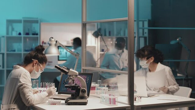 Timelapse of young female chemist in lab coat, protective mask, gloves and eyewear carrying out clinical research by workplace equipped with microscope, laptop and test tubes containing liquids