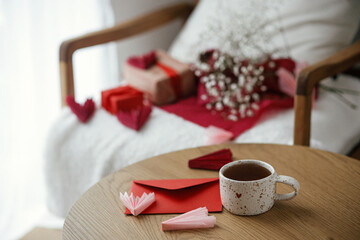 Happy valentines day! Cute modern cup of tea with heart on wooden table against stylish chair with gifts, red envelope and white flowers. Valentine morning surprise for beloved. Space for text