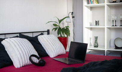 Laptop with black blank screen and headphones on bed with viva magenta bed sheet, black blanket and black and white pillows in modern apartment. Freelancer, blogger, business outsourcing. Home office