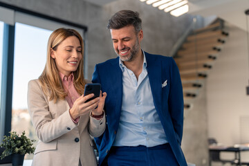 Joyful employers business man and woman friends at work in suits looking at smartphone, using mobile apps tech in office during the pause from work.