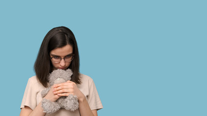 Sad beautiful student girl with glasses, hugging a gray teddy bear on a blue background