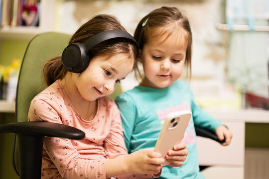 Two little girls sisters wear headphones watching cartoons or kid video on mobile phone.
