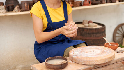 hands of craft woman working with clay lump in pottery studio outdoors in village on background of shelves with ready-made clay products