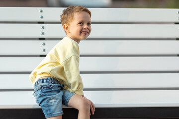 A happy little boy sits on a bench in a city park on a sunny day © Irina Mikhailichenko