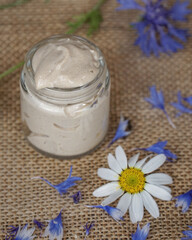 Natural homemade eye cream in transparent glass jar with cornflowers and chamomile flowers on the beige, white, blurred background.