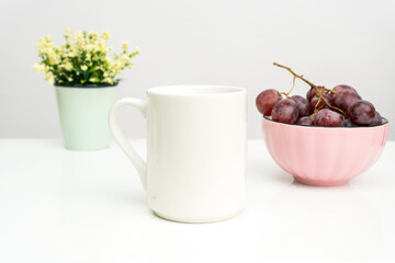 A white blank tea mug on the top of a white table with the grapes in a bowl placed behind it