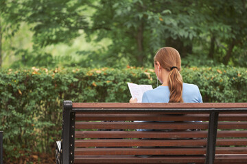 Fototapeta premium A young woman is sitting on a park bench, studying the price list of amusement rides. She has a cell phone earpiece in her ear.