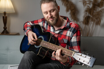 Obraz premium Happy young man playing the guitar while sitting on sofa at home in the evening, enjoying carefree time at home.