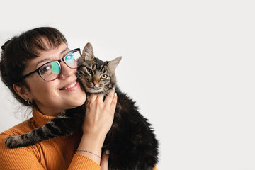 Latin woman wearing eyeglasses is holding her tabby cat while smiling and looking at camera....