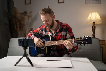 Male blogger teaches guitar playing in an online lesson at home.