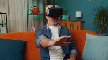Bearded senior man using virtual reality futuristic technology VR app headset helmet to play simulation 3D 360 video game, watching film movie at modern home apartment. Guy in goggles sitting on sofa