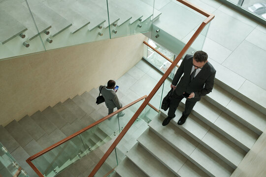 Above shot of two intercultural businessmen in formalwear using mobile phones while walking downstairs inside modern office center