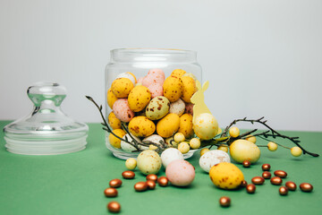 Chocolate colorful decorative eggs in a glass jar on a white-green background. Happy Easter. Front view
