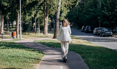 Beautiful young woman in white suit is riding to work on her electric scooter in city parkland