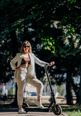Beautiful young woman in sunglasses and white suit is standing with her electric scooter in city parkland