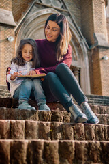 Mother and daughter sitting at the stairs and read a book outdoor. Low angle view. 