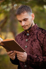 Fototapeta premium A young man in medieval brown clothes stands with an old book in his hand. A guy in a historical costume in nature.