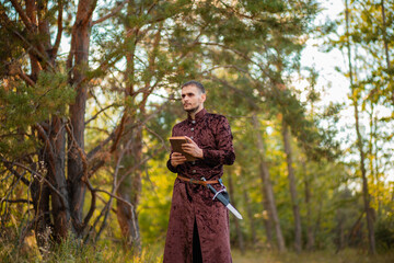 A young man in medieval brown clothes stands with an old book in his hand. A guy in a historical costume in nature.