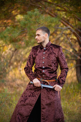 A young man in medieval brown clothes stands in the forest with a dagger. A guy in a historical costume in nature.