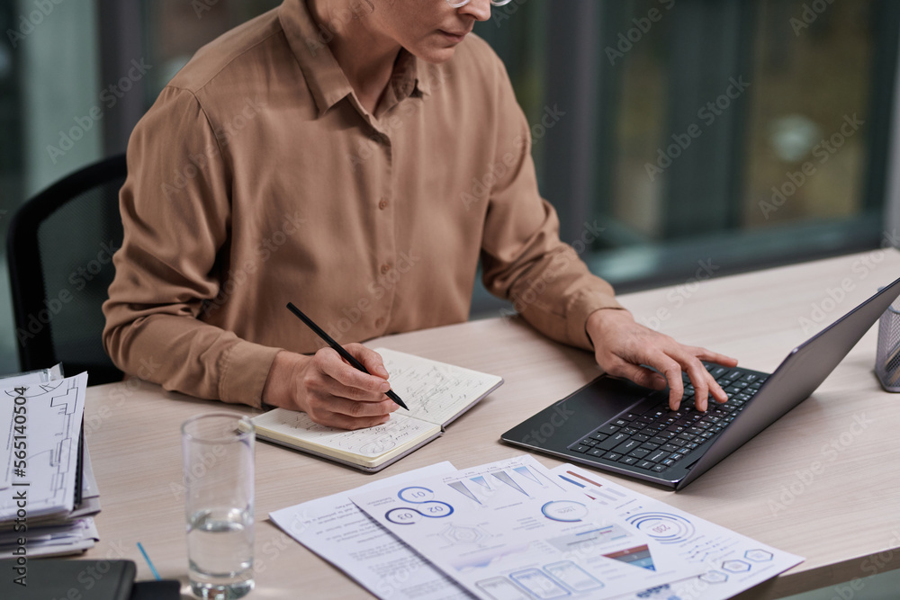 Poster Cropped shot of experienced female broker or analyst with laptop networking and making notes in notebook by workplace in office