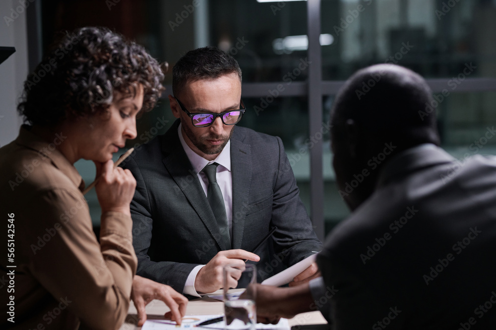 Leinwandbilder focus on serious broker analyzing financial document in front of two intercultural colleagues discus
