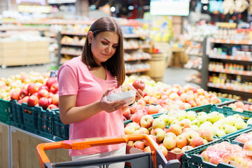 Young brunette woman picks chooses fruits on the counter in supermarket. Female housewife shopping in market standing near vegetable department store with a grocery cart.