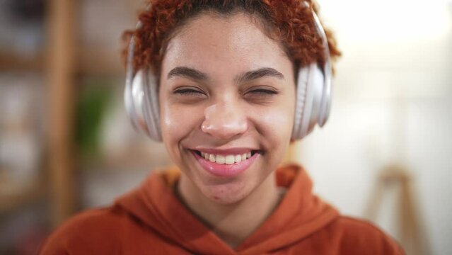 Portrait face of beautiful african american young woman, sits at home on bed in headphones and listens to music. Enjoy listening to favorite tracks. Happy toothy smile and laugh. Good mood and rest.