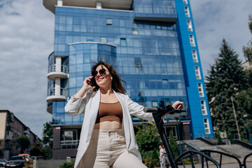 Beautiful young woman in sunglasses and white suit standing on her electric scooter near modern building and talking phone