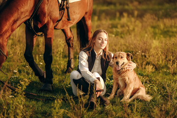 A young beautiful woman jockey with her dog sits in a meadow near her horse at sunset.