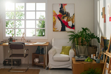 Workplace of modern artist by window of spacious home studio with table, chair, armchair, abstract painting on wall and green plants