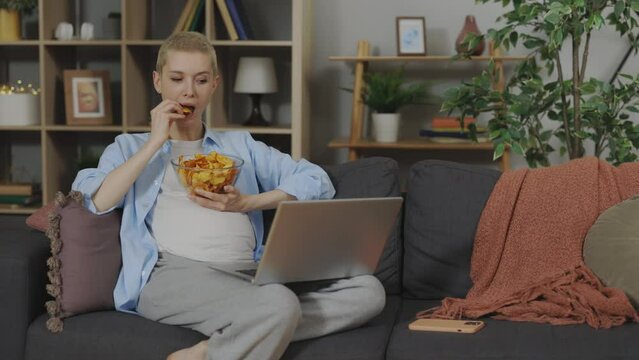 Attractive Pregnant Woman Eating Potato Chips And Watching New Series On Laptop While Sitting On Couch At Home. Unhealthy Junk Food During Pregnancy.