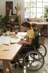Adolescent girl in wheelchair sitting by wooden table with supplies for art creation in her home studio and drawing new masterpiece