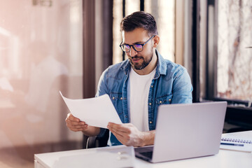 Satisfied young man with glasses sitting at a desk and doing paperwork at his workplace.