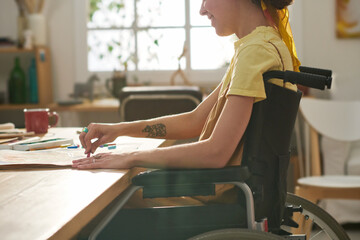 Close-up of happy young woman with disability sitting in wheelchair by table in studio and drawing with crayon on page of notepad