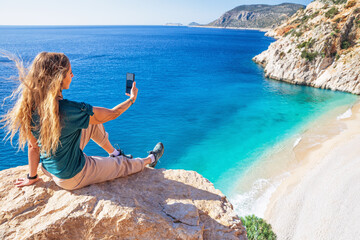 Young woman making selfie photo by smartphone over Kaputas beach, Lycia coast. Summer day walk by Lycian way at family vacation in Mediterranean Sea, Kas, Antalya region, Turkey - obrazy, fototapety, plakaty
