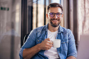 Handsome smiling young man drinking coffee sitting at office desk and looking at camera