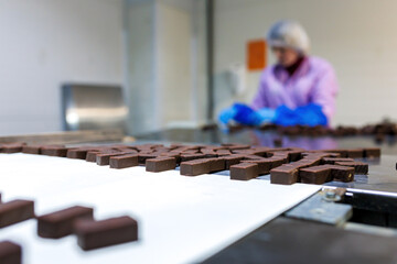 Professional female worker in uniform and protective gloves sorts chocolate candies on production line at factory - 565154991