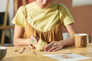 Close-up of young creative craftswoman making human face out of piece of clay while sitting by workplace with all necessary supplies