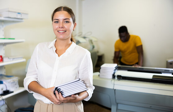 Portrait of friendly female employee of printing house with a stack of notebooks in her hands