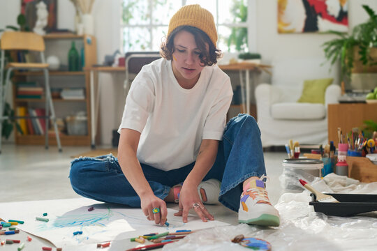 Young female artist or designer with blue crayon drawing on paper while sitting on the floor of spacious and comfortable home studio