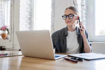 A woman talking on the phone in the office in a shirt, her hair neatly removed, glasses eyes, an online designer working workplace. Coworking space using a laptop computer.