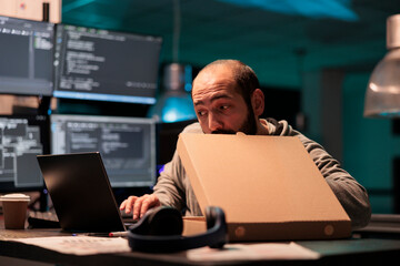Software developer eating pizza from takeaway delivery, taking bite of food while he works after...