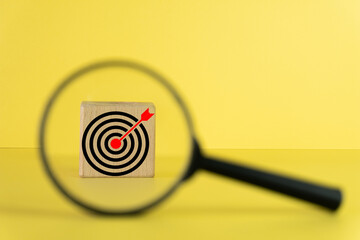 A magnifying glass focus on a wooden cube with target board icon over yellow background use for business,analysis,attention to detail,education,knowledge, research idea.