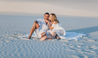 The bearded man and the blonde girl are hugging while sitting on the white sand and enjoying the sunset. Desert and newlyweds. Love story of cheerful and loving people. Vacation, honeymoon. - Powered by Adobe