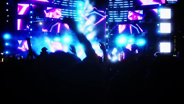 People jump and dance at the evening concert with light show.Fans raise their hands in front bright colorful strobe lights on stage. Crowd is dancing, filming performance rock band, throwing party. 4K