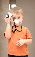 Fototapeta premium People. Handsome funny and emotional caucasian blond little boy in an orange t-shirt with a children's white camera takes pictures in the home interior.