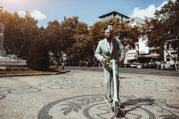 View of a fancy bald beard mature black guy riding an e-scooter over the paving stone; an elegant African businessman in a green tailored suit is riding an electric scooter of the same green color