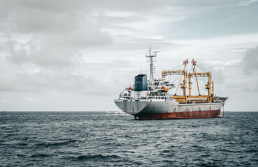 View from behind of a huge freighter in the ocean with two cargo cranes on it; a big cargo ship in open waters with a copy space place on the left; an overcast skyscape in the background